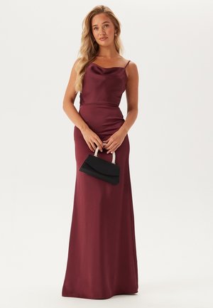 Abito da sera in satin color vino con scollatura a cappuccio e spalline sottili. Il modello tiene una pochette nera con manico di perline.