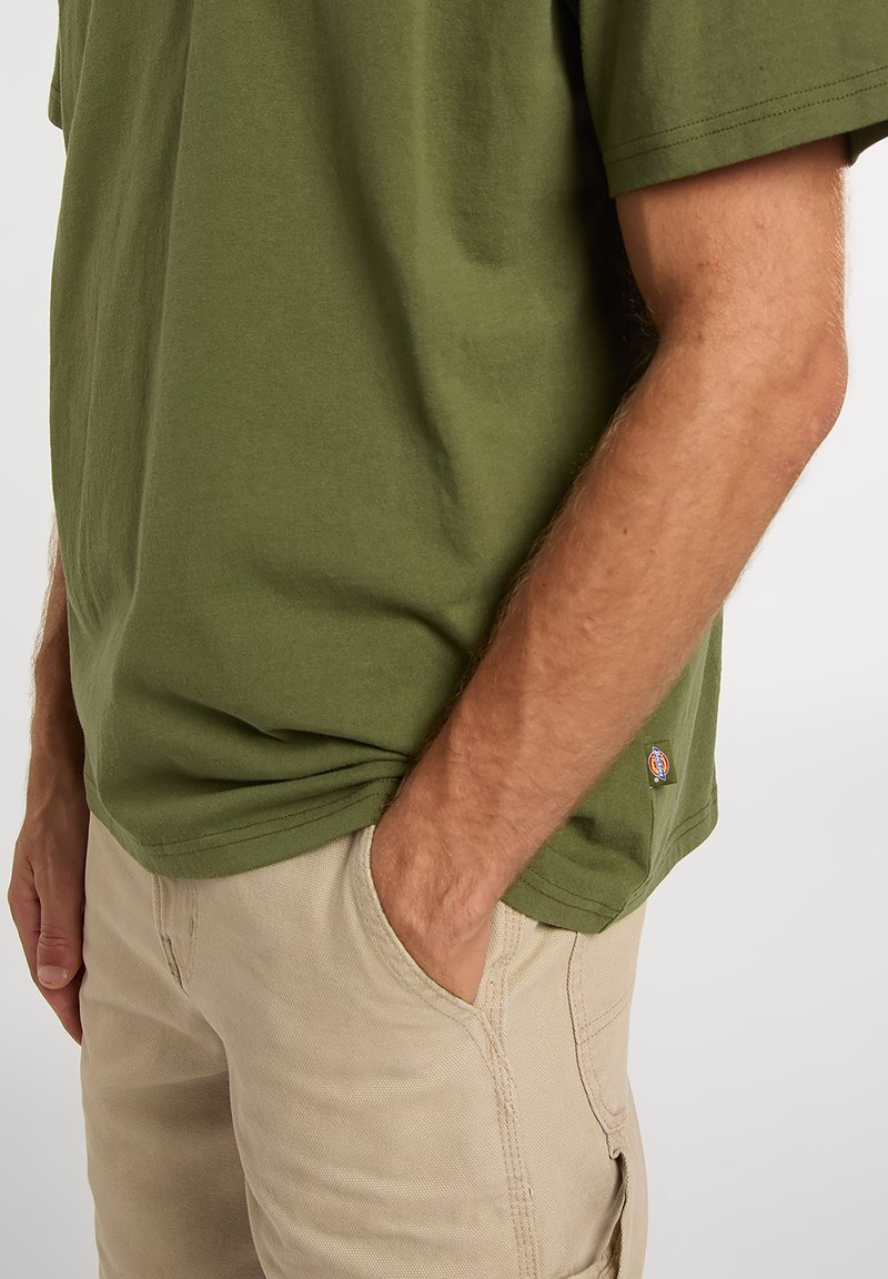 T-shirt en coton vert olive à manches courtes, coupe ample, avec un petit patch logo. Short beige avec poches, détails de coutures visibles.