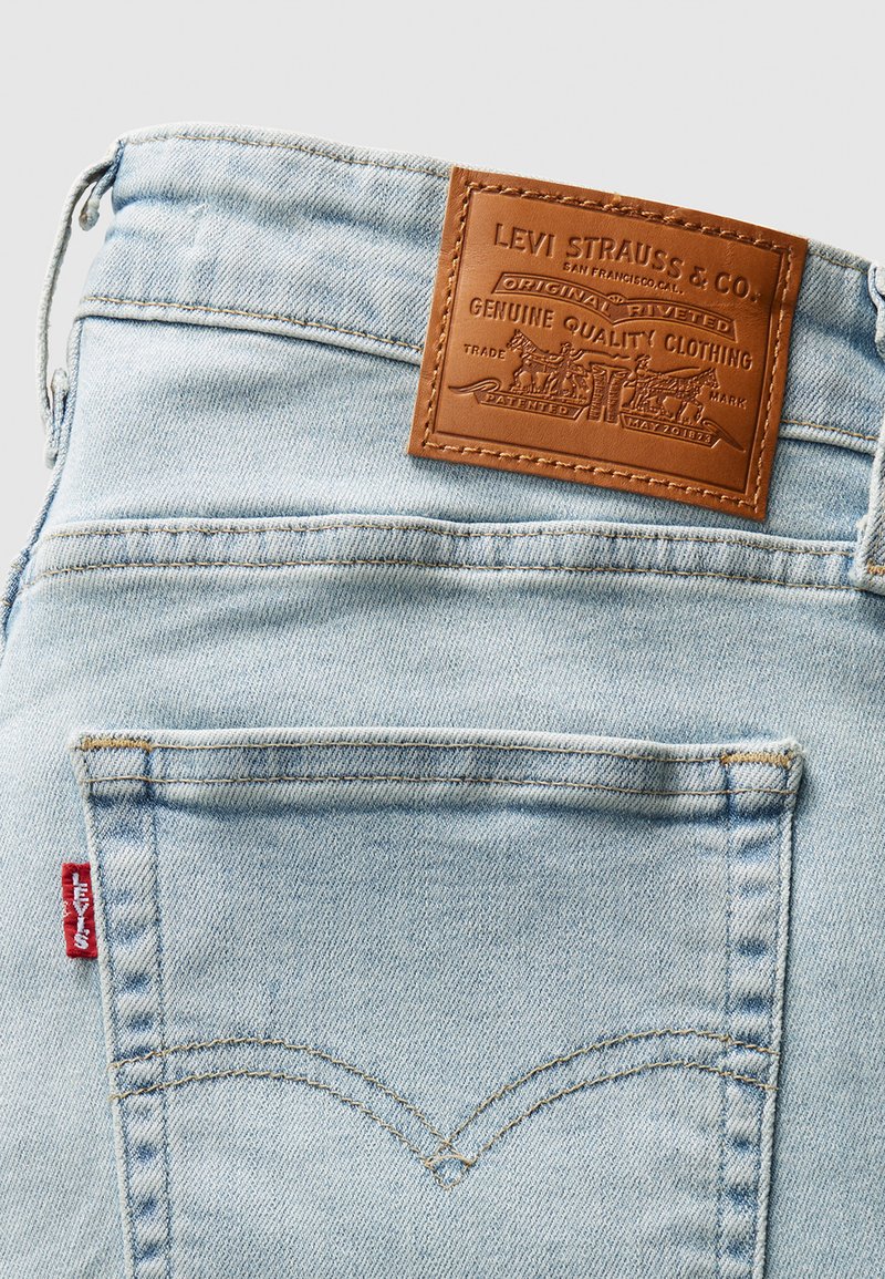 Jeans de mezclilla azul claro con un parche de cuero con logotipo en relieve. Bolsillo trasero destacado con costura amarilla y detalle de etiqueta roja.