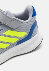 Baskets de sport grises avec tige en mesh, accents jaunes vifs, détail de talon bleu et semelle blanche avec un motif texturé pour une meilleure adhérence.