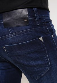 Jeans de mezclilla azul oscuro con un parche de cuero etiquetado "ANTONY MORATO". Presentan detalles metálicos y un diseño clásico de cinco bolsillos.