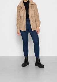 Giacca in peluche beige con colletto, tasche frontali e chiusura a bottoni, abbinata a jeans attillati blu scuro e stivaletti neri con tacco chunky.