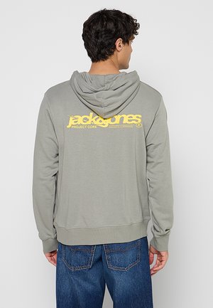 Férfi szürke kapucnis pulóvert visel hátul sárga „Jack & Jones Project Core” logóval, kék farmerrel párosítva, háttal néz.