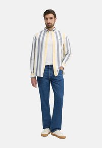 Camisa de botones a rayas en gris, amarillo y blanco, superpuesta a una camiseta blanca, combinada con vaqueros azules y zapatillas beige.