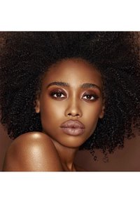 Close-up van een gezicht met donker, krullend haar en een egale teint. Kenmerkt zich door warm getinte make-up, waaronder bronzen oogschaduw en een nude lipkleur.