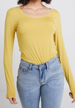 Kobieta ubrana w dopasowany, musztardowo-żółty, długi rękaw z koronkowym wykończeniem na dekolcie i mankietach, w połączeniu z jasnoniebieskimi, wysokim stanem jeansami.