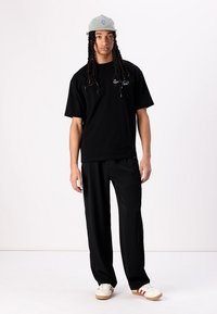 Svart bomulls t-skjorte med korte ermer og brodert tekst, kombinert med svarte bukser og kremfargede sneakers med røde striper. Lysfarget caps.