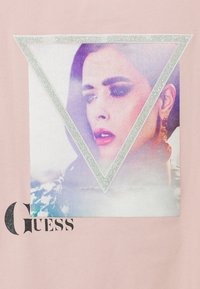 Rožinė marškinėliai su spausdintu trikampiu grafiku, vaizduojančiu moters veidą, su blizgučiais ir prekės ženklu "Guess" juoda spalva.