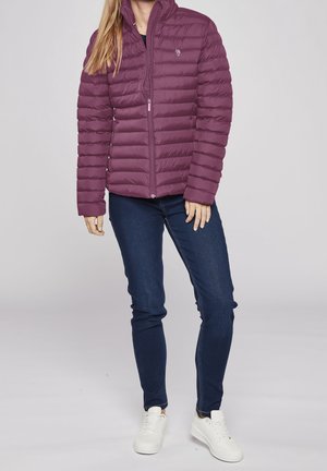 Femme portant une veste matelassée violette zippée, un jean skinny bleu foncé et des baskets blanches, debout devant un fond uni.