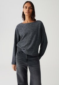 Femme aux longs cheveux noirs portant un haut à manches longues texturé gris charbonneux et un jean taille haute gris foncé, sur un fond clair uniforme.