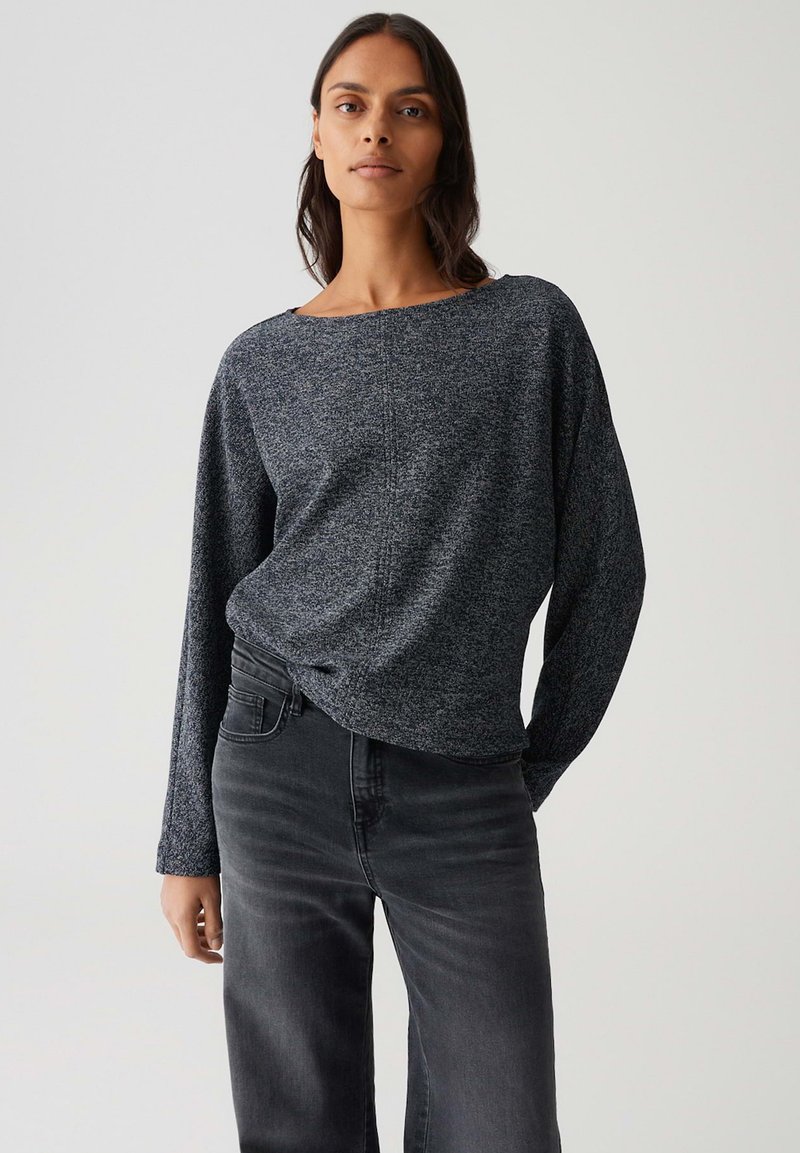 Femme aux longs cheveux noirs portant un haut à manches longues texturé gris charbonneux et un jean taille haute gris foncé, sur un fond clair uniforme.