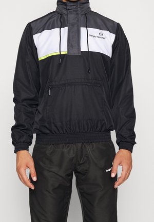 Man wearing black Sergio Tacchini tracksuit with white and neon yellow stripe on jacket and elastic cuffs.