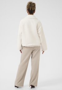 Cream textured jacket with a collar and wide sleeves, worn over beige wide-leg trousers. White background highlights the outfit's color contrast.