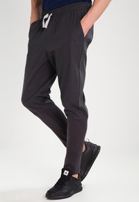 Svarta sportbyxor med en avsmalnande design, elastisk midja och vit snörning, kombinerade med svarta sneakers. Slät textur, inga mönster.