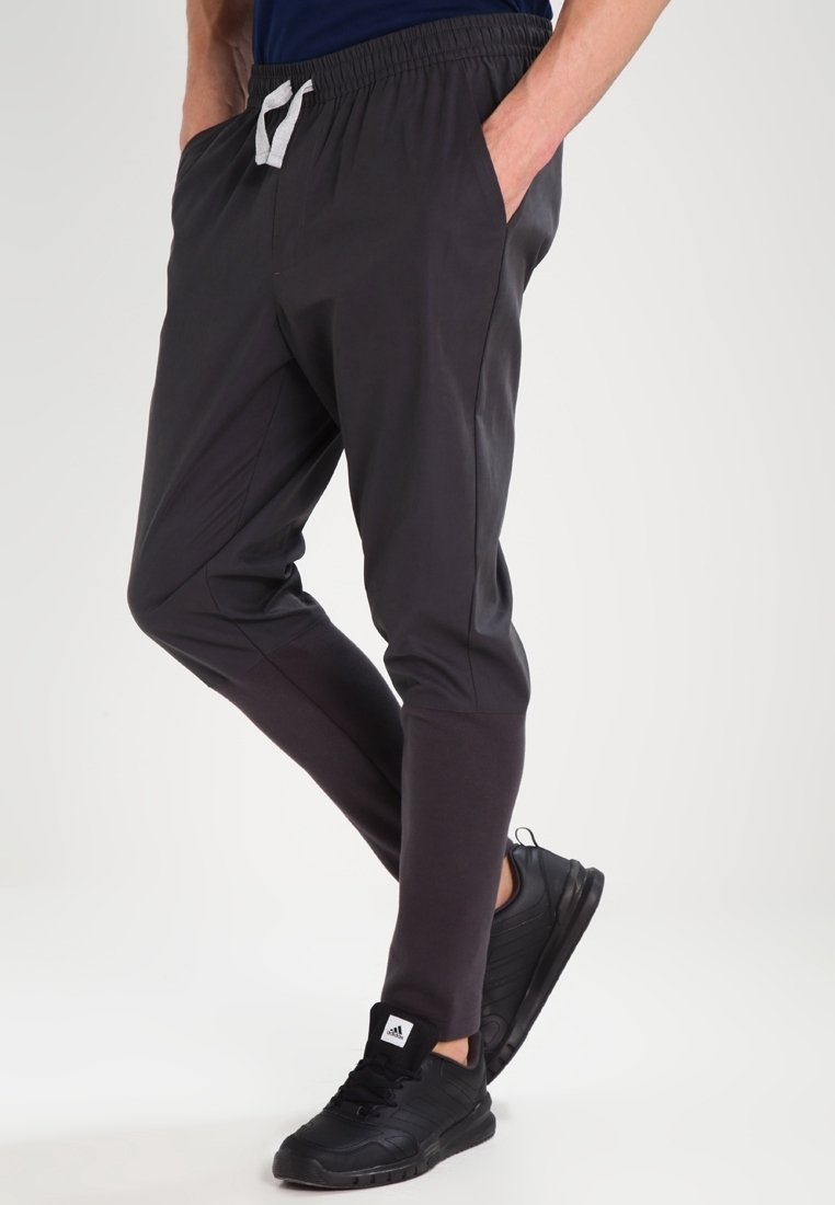 Svarta sportbyxor med en avsmalnande design, elastisk midja och vit snörning, kombinerade med svarta sneakers. Slät textur, inga mönster.