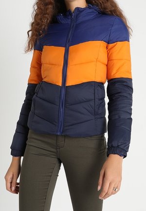 Femme portant une doudoune bicolore bleu marine et orange avec une fermeture éclair frontale et des manches longues, associée à un pantalon vert olive.