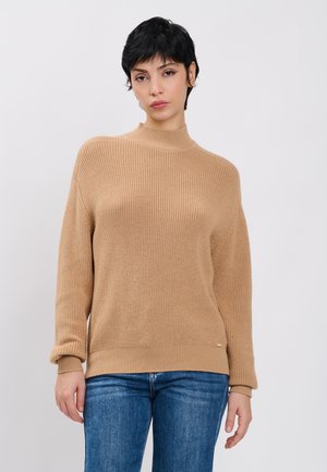 Kvinde med kort sort hår iført en beige striktrøje og blå jeans, stående mod en ensartet lys grå baggrund.