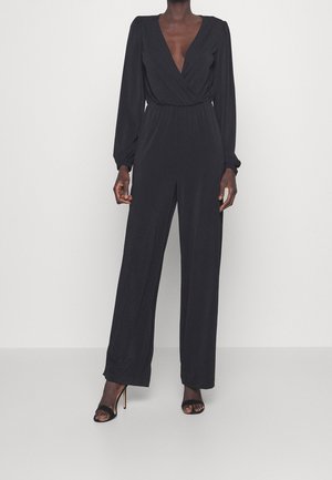 Zwarte jumpsuit met lange mouwen, diepe V-hals, wikkeldesign, elastische tailleband, vloeiende stof en brede pijpen, gecombineerd met openhiel sandalen.