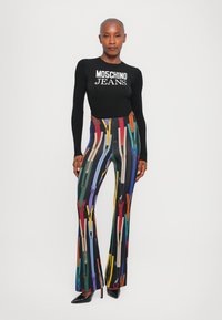 Čierny rebrovaný top s dlhými rukávmi s logom "MOSCHINO JEANS", doplnený farebnými rozšírenými nohavicami s vzorom zipsov. Čierne vysoké topánky.