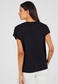 Zwarte katoenen t-shirt met korte opgerolde mouwen, ronde hals en rechte snit, weergegeven van achteren op een model.