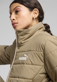 Kreemikattega vooderdatud jakk kõrge kraega, millel peal valge Puma logo vasakul rinnal, lukuga kinnitamine ja sile tekstuur.