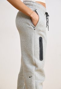 Pantalon de jogging gris avec taille élastique et cordon de serrage, présentant une poche zippée sur le côté et un petit logo noir près de la cheville.