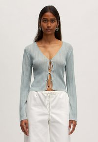 Hellblau gestreifter Pullover mit tiefem V-Ausschnitt und Knotenfrontverschluss. Lange Ärmel und ein leicht verkürzter Schnitt. Kombiniert mit weißen Hosen.