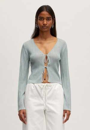 Light blue ribbed sweater with a deep V-neck and tie-front closure. Long sleeves and a slightly cropped design. Paired with white trousers.
