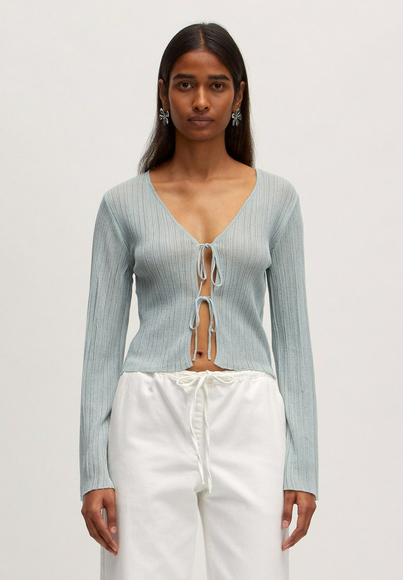 Hellblau gestreifter Pullover mit tiefem V-Ausschnitt und Knotenfrontverschluss. Lange Ärmel und ein leicht verkürzter Schnitt. Kombiniert mit weißen Hosen.