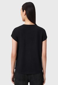 Γυναίκα με μακριά σκούρα μαλλιά, που φοράει μια απλή μαύρη κοντομάνικη μπλούζα και σκούρο τζιν παντελόνι, στέκεται με την πλάτη προς την κάμερα σε φωτεινό φόντο.