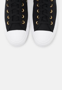 Mustat kangaskengät valkoisilla kumipohjilla, joissa on kultaiset nauhaku complexity ja mustat nauhat, esitetty ylhäältä alas näkökulmasta.