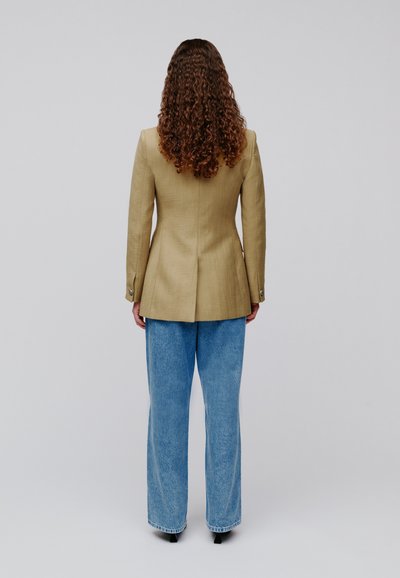 Άτομο με μακριά σγουρά μαλλιά που φοράει ταμπά σακάκι και άνετο μπλε τζιν, στέκεται με την πλάτη προς τα εμπρός σε ένα απλό ανοιχτό γκρι φόντο.