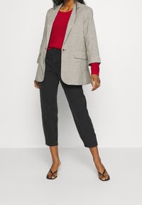 Blazer a scacchi in beige e nero, indossato sopra un maglione rosso. Jeans neri a vita alta, abbinati a infradito neri, evidenziando un look rilassato.