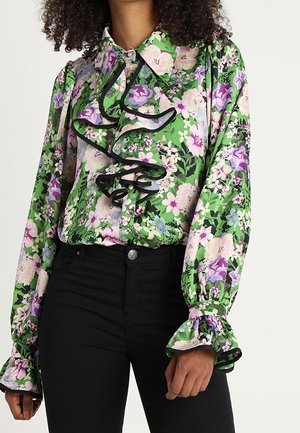 Vrouw draagt een groene blouse met bloemenpatroon in paars en roze, zwarte ruches, opgeblazen mouwen en zwarte, nauwsluitende broek.