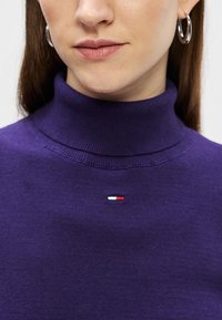 Fioletowy sweter z golfem wykonany z dzianiny. Posiada małe logo marki z czerwonymi, białymi i niebieskimi paskami na piersi.