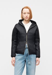 Νεαρό άτομο με κοντά κόκκινα μαλλιά, φοράει μαύρο καπιτονέ μπουφάν με κουκούλα πάνω από λευκό πουκάμισο και ανοιχτόχρωμα τζιν μπροστά σε λευκό φόντο.