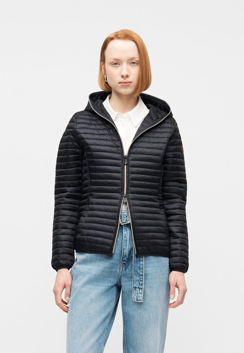 Νεαρό άτομο με κοντά κόκκινα μαλλιά, φοράει μαύρο καπιτονέ μπουφάν με κουκούλα πάνω από λευκό πουκάμισο και ανοιχτόχρωμα τζιν μπροστά σε λευκό φόντο.