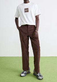 Bílý tričko s tmavohnědým logem "HUF", doplněné hnědými kordovými kalhotami a šedými běžeckými botami se žlutými akcenty.