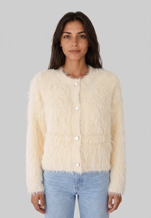 Donna che indossa un cardigan beige chiaro peloso con bottoni e jeans azzurri, in piedi davanti a uno sfondo grigio uniforme.