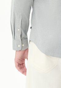 Grijze langlopende shirt met knoopsluiting bij de manchetten, met een gladde textuur en een gebogen zoom, gecombineerd met lichte stof rond de taille.