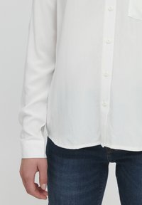 Hvid skjorte med knapper, lavet af glat stof, der har en let glans, lange ærmer og en afrundet kant, kombineret med mørkeblå jeans.