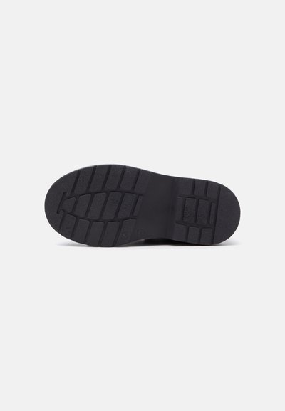 Semelle de chaussure noire avec des rainures profondes rectangulaires et carrées disposées pour la traction et la stabilité, vue de dessous.