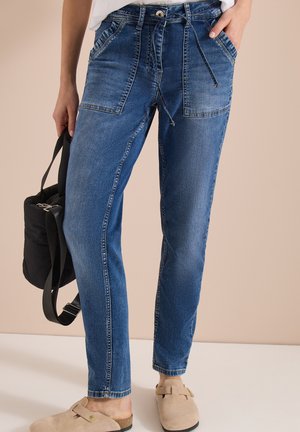 Personne portant un jean en denim bleu avec de grandes poches à l'avant, des chaussures beiges sans lacets, tenant un sac à dos noir à côté d'elle.