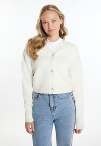 Weiße, verkürzte Strickjacke mit V-Ausschnitt, fünf Knöpfen auf der Vorderseite und gerippten Bündchen. Kombiniert mit hellblauen Jeans.