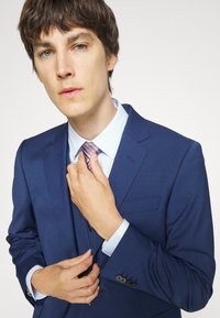 Sacou ajustat de culoare bleumarin, cu الأ lapels notch, mâneci lungi și trei butoane. Cămașă de birou de culoare bleu deschis și cravată cu dungi. Finisaj din țesătură netedă.