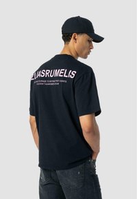 Mies, który ma na sobie czarną czapkę, czarny t-shirt z dużym ró�żowym napisem z tyłu oraz ciemne, zniszczone dżinsy, stojący bokiem z rękami w kieszeniach, na jasnoszarym tle.
