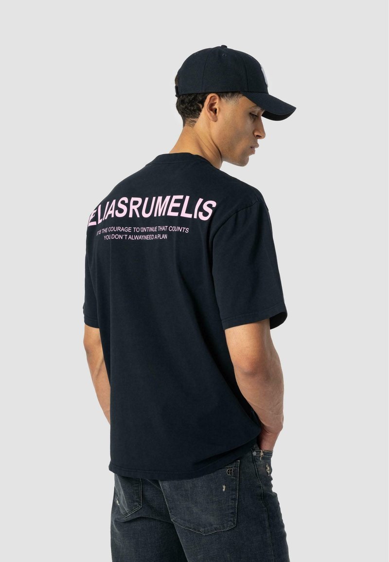 Mies, który ma na sobie czarną czapkę, czarny t-shirt z dużym różowym napisem z tyłu oraz ciemne, zniszczone dżinsy, stojący bokiem z rękami w kieszeniach, na jasnoszarym tle.