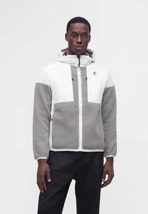Hombre con chaqueta de forro polar blanca y gris con capucha y cremallera frontal, y pantalones negros, de pie frente a un fondo blanco liso.