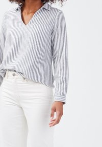 Chemise à rayures à manches longues en coton gris et blanc, avec un col en V, une poche et des accents circulaires subtils, associée à un jean blanc.