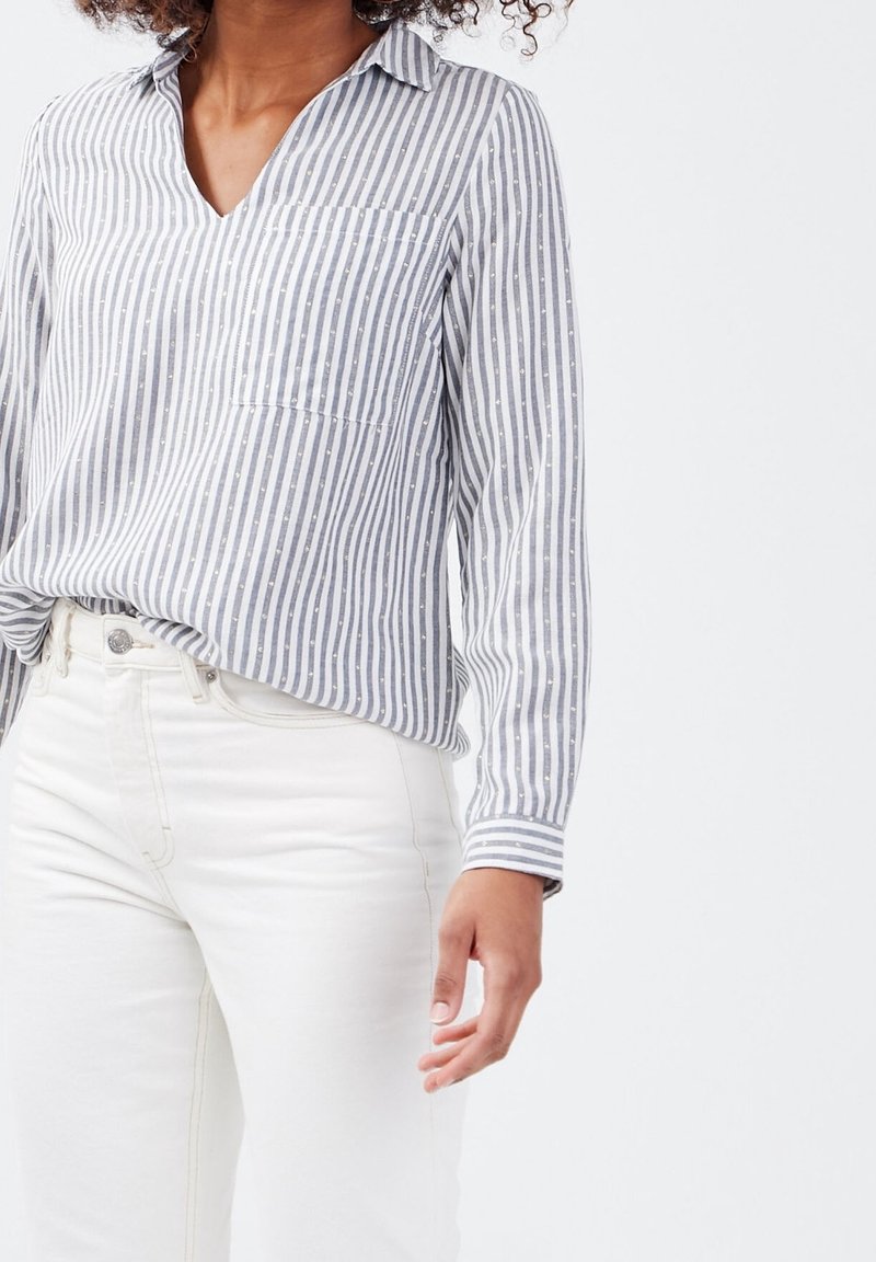 Chemise à rayures à manches longues en coton gris et blanc, avec un col en V, une poche et des accents circulaires subtils, associée à un jean blanc.